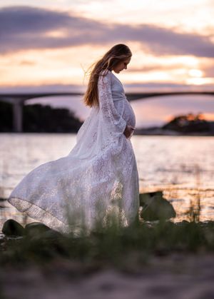 Pregnant woman in a white lace gown holds her belly by a riverside at a stunning pink and purple sunset.