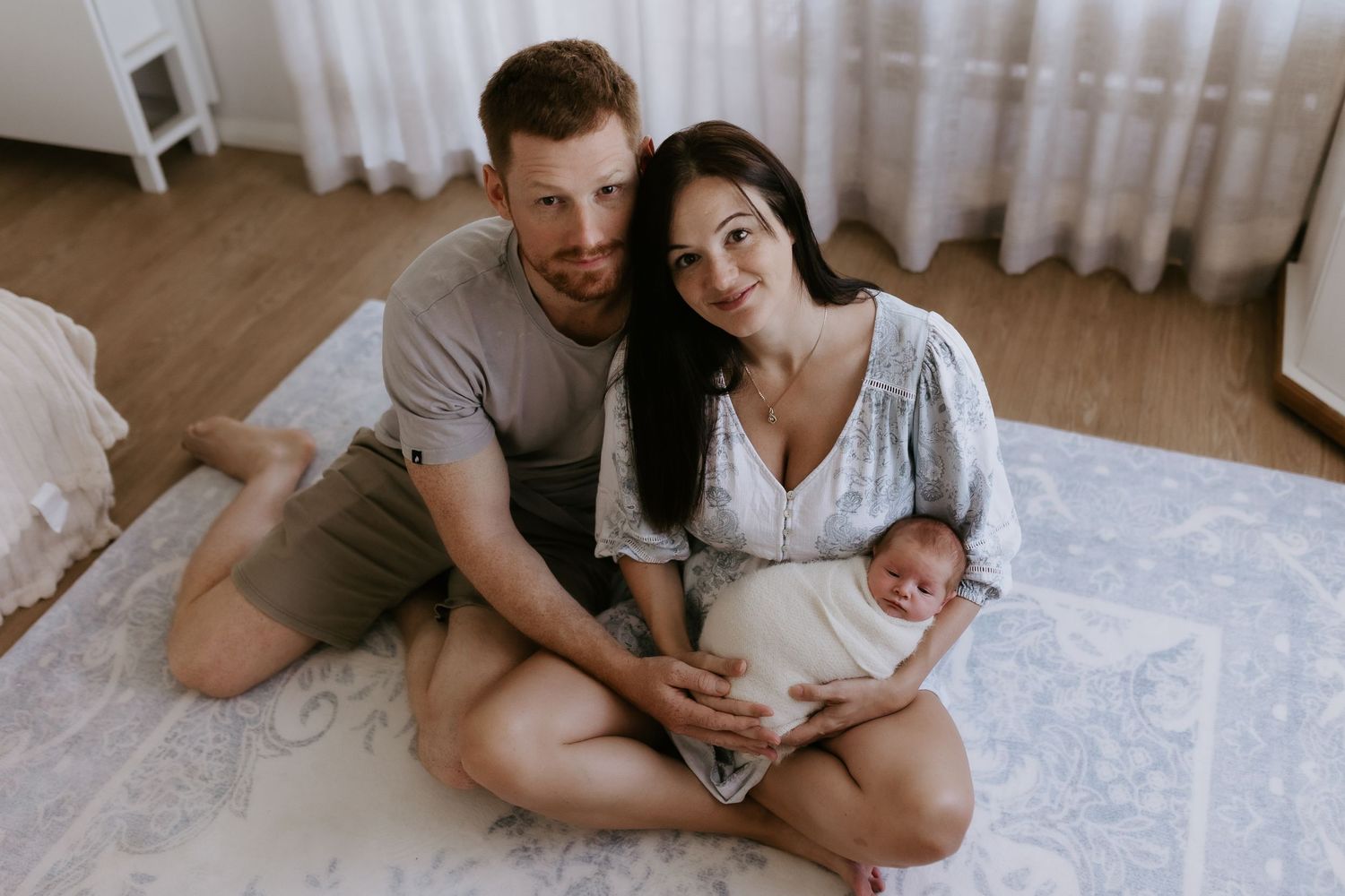 A couple sits on a floor holding their swaddled newborn baby in a bright, cozy nursery room.