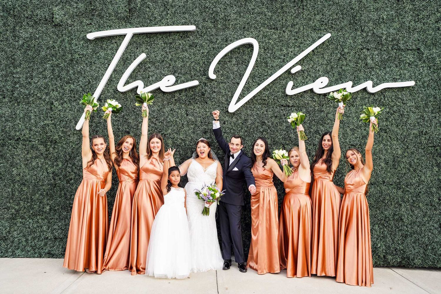 Bride and groom celebrate with bridesmaids in coral satin gowns holding bouquets in front of The View venue