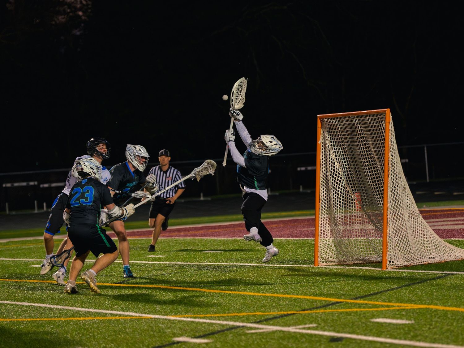 Janesville lacrosse goalie leaps with stick raised for a save near the net under the lights as defenders and a referee track the ball.