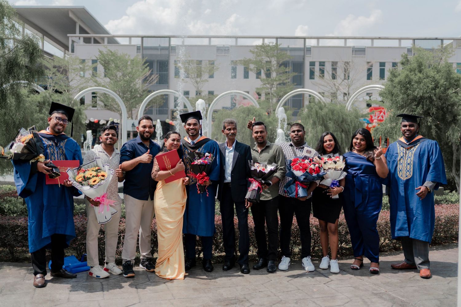 Group of graduates in blue robes celebrating outdoors, holding flowers after university graduation ceremony.