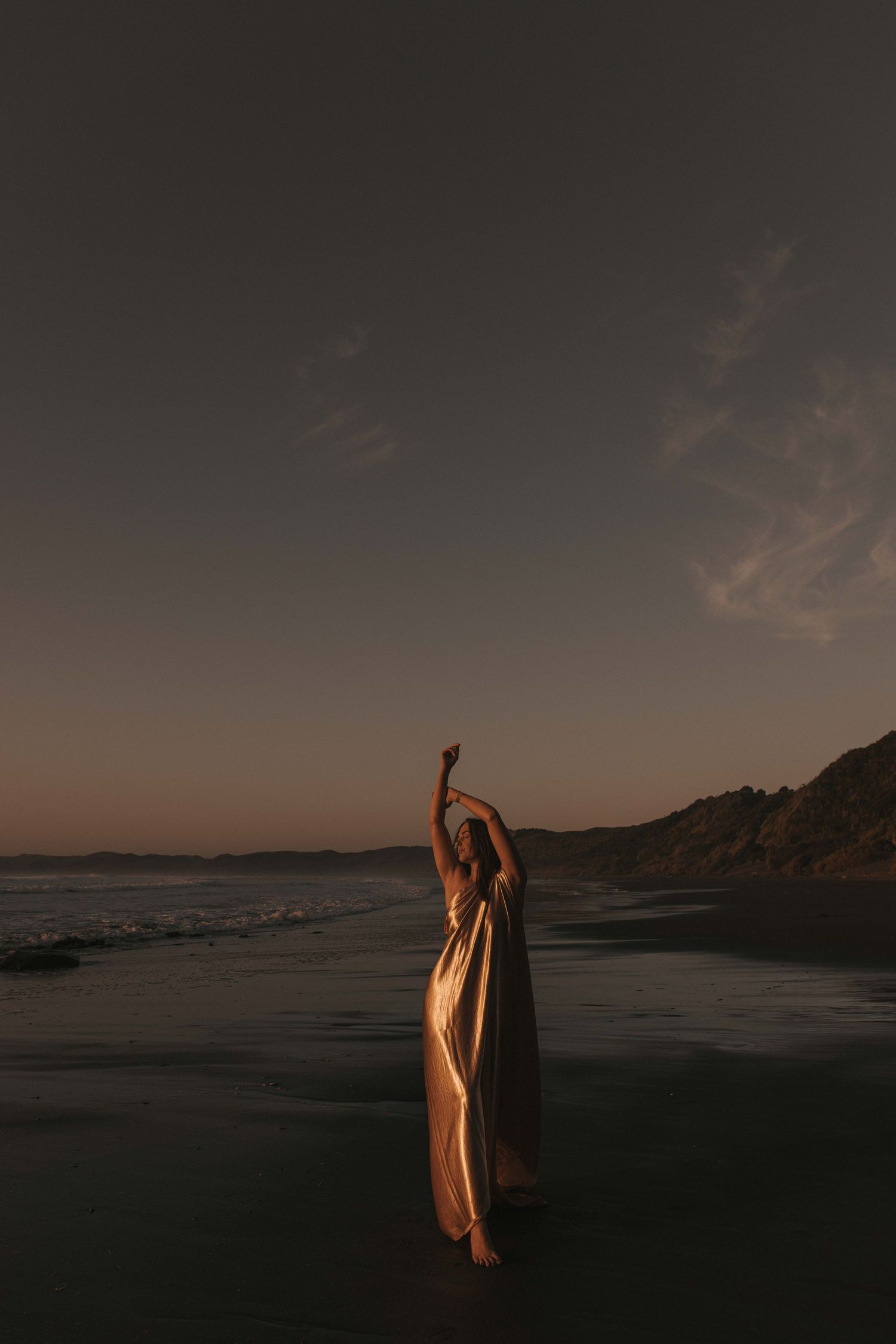 A woman in a flowing dress stands on a beach at dusk, arms raised gracefully toward the twilight sky.
