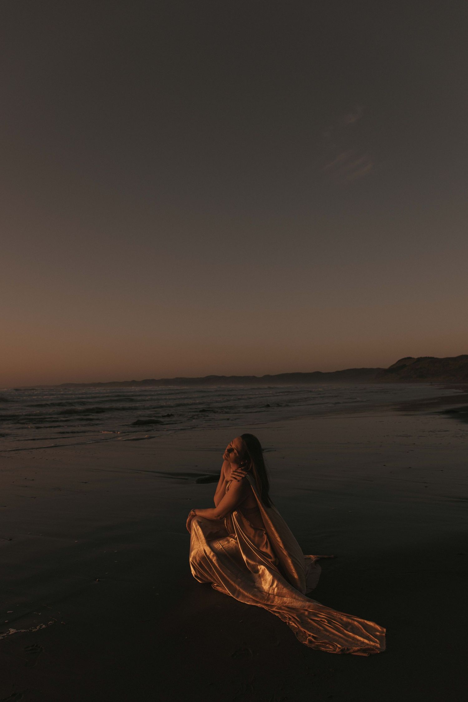 Woman draped in gold fabric on Raglan's black sand beach at sunset — Goddess Shoot® by Nami Creative