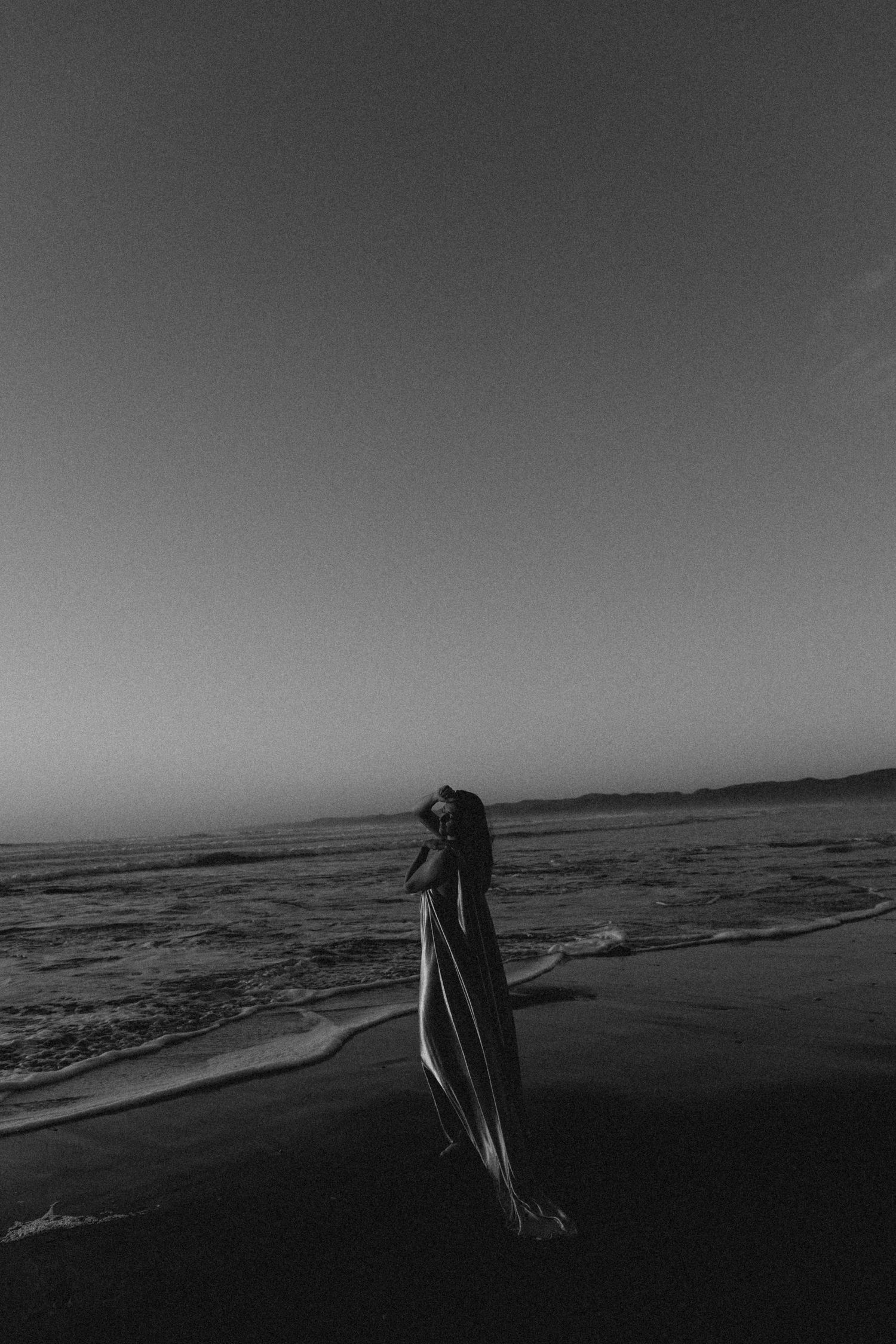 Black and white photo of a woman in a flowing dress standing on a dark beach at dusk with ocean waves.