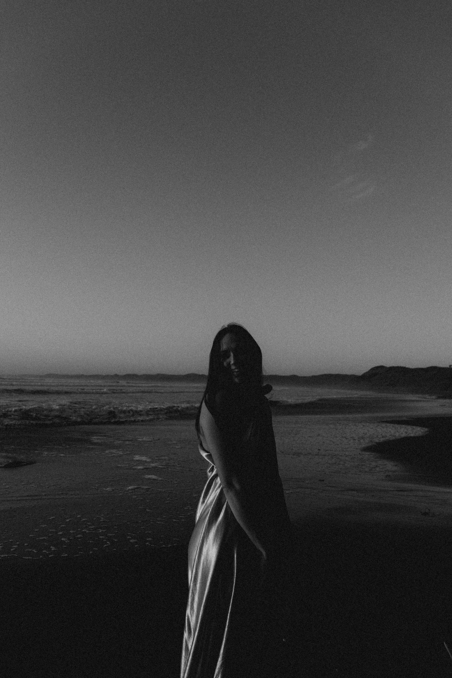 Black and white photo of a woman wrapped in fabric standing on a dark beach at dusk with ocean waves behind her.