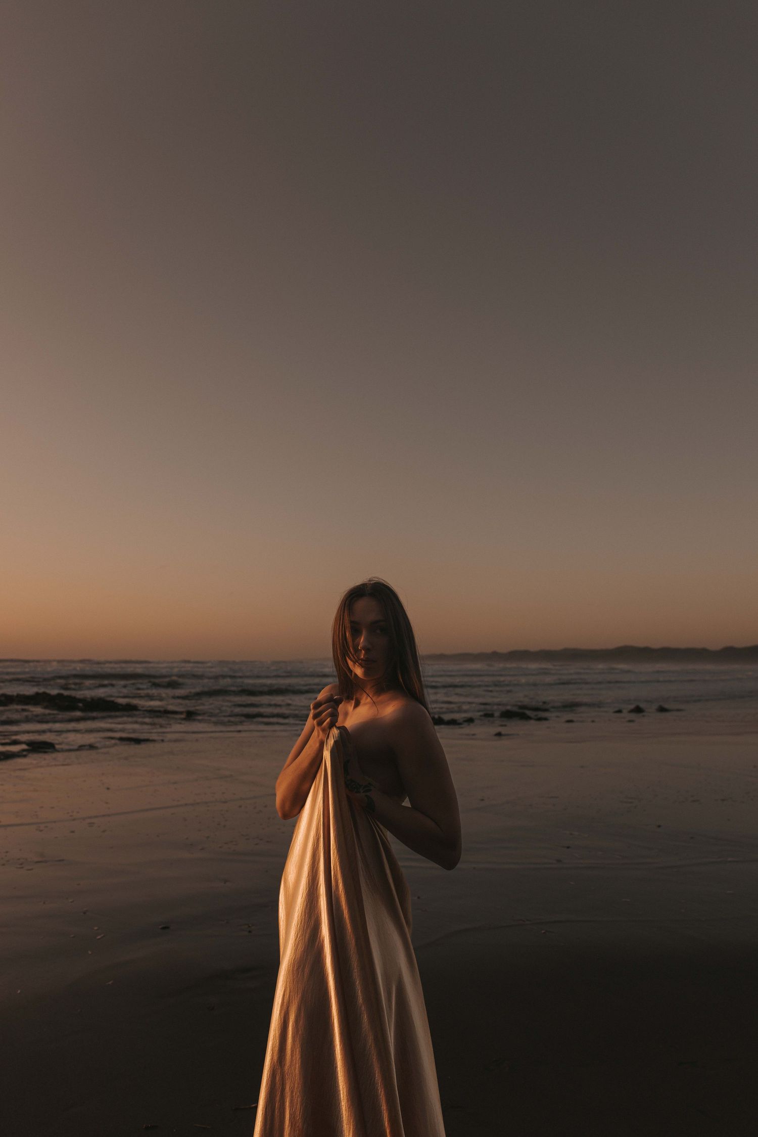Woman in white dress standing on beach at sunset with ocean waves and golden sky in background.