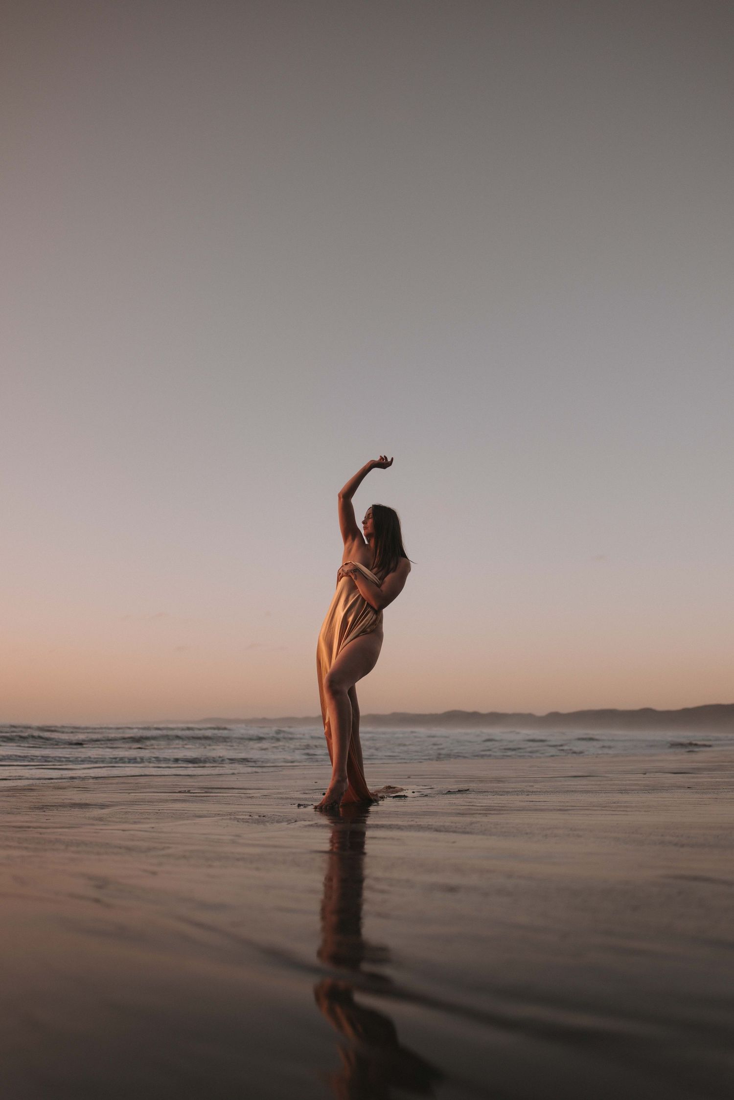 A woman in a flowing dress poses gracefully on a reflective beach at sunset with arms raised.