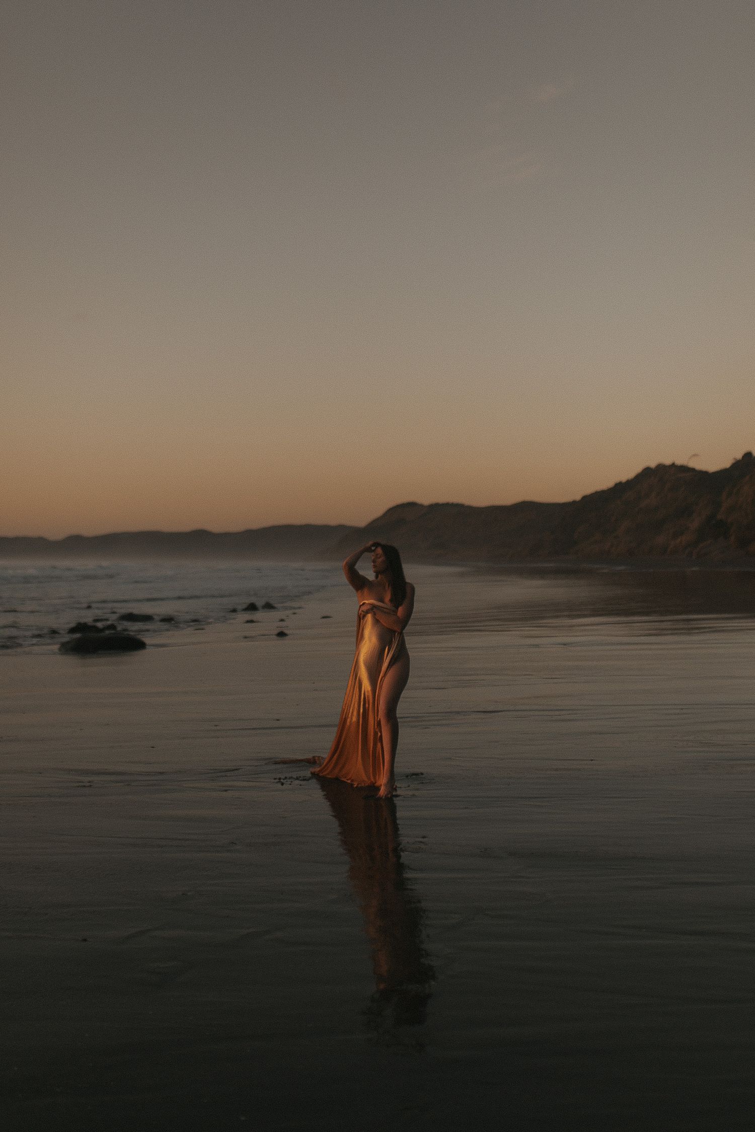 Woman draped in gold fabric on Raglan's black sand beach at sunset — Goddess Shoot® by Nami Creative