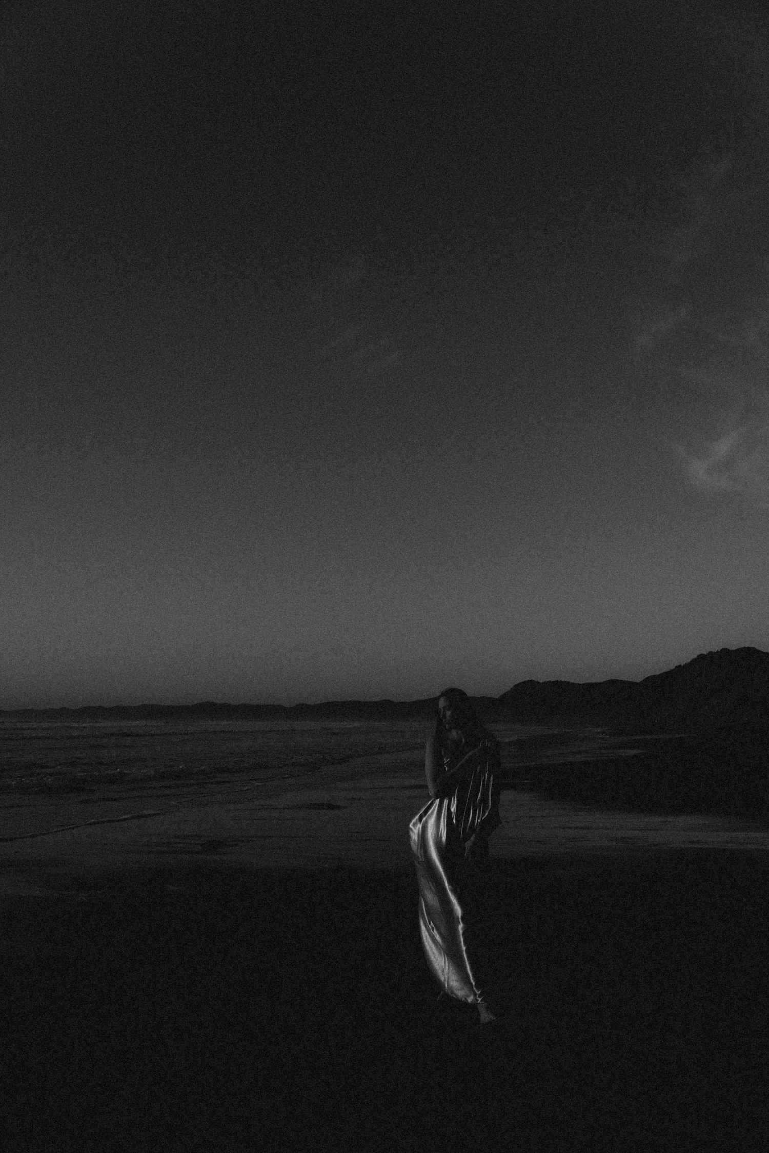 A lone figure in a flowing dress stands on a dark beach at night in this moody black and white photograph.