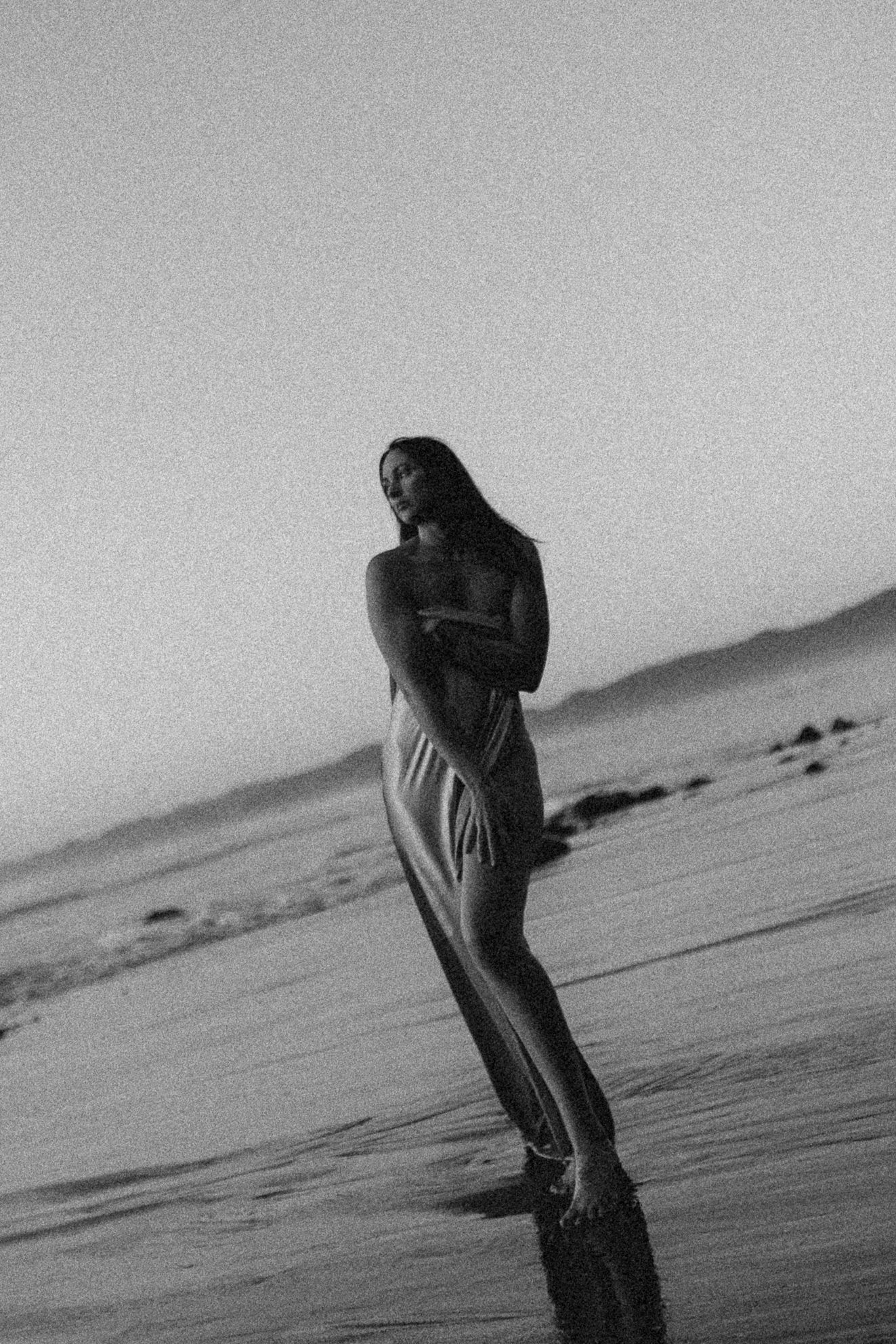 Black and white photo of a woman in a long dress standing on a beach at dusk.