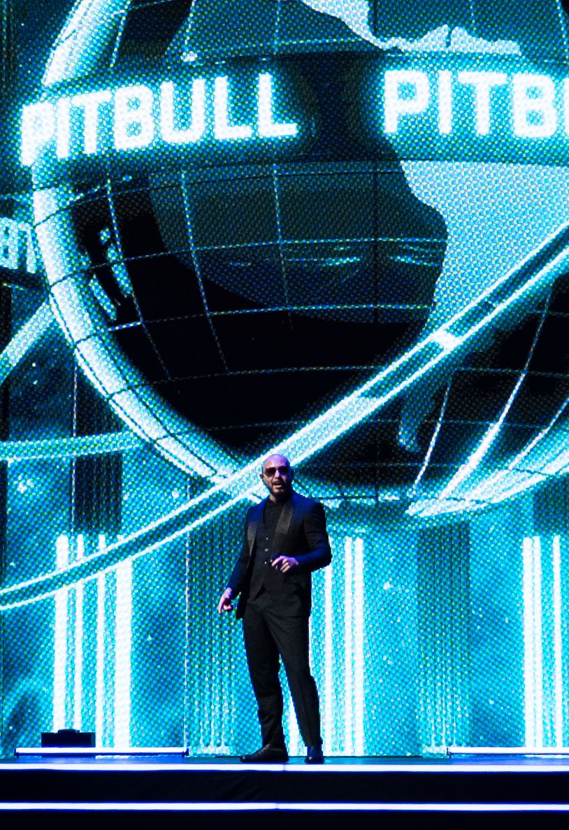 Pitbull performing on stage with glowing blue globe backdrop and neon lights at a live concert event.