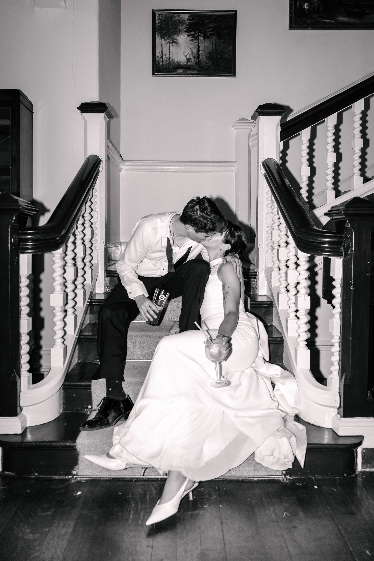 Black and white photo of bride and groom kissing on elegant staircase, holding cocktails, bride in white gown.