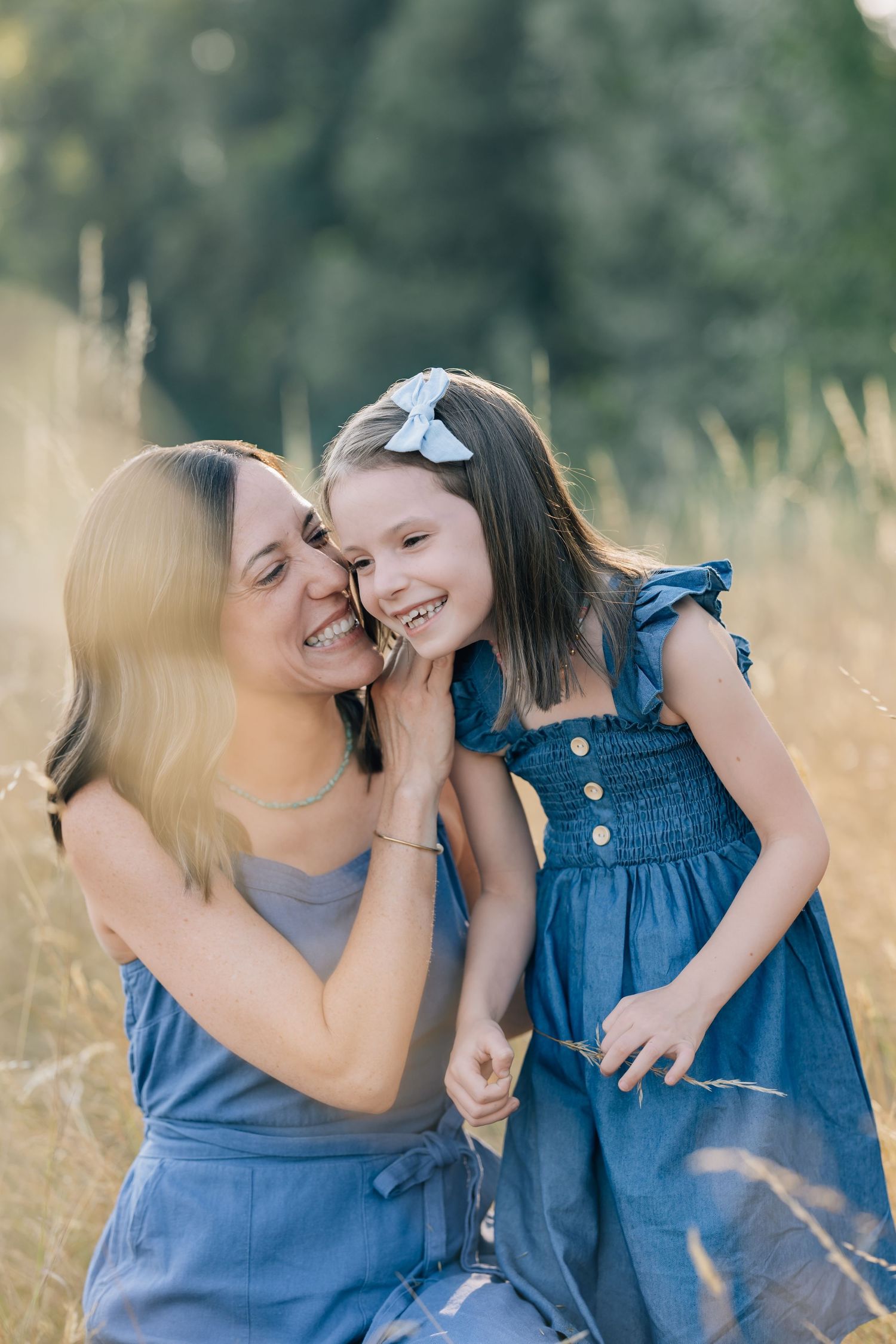A mother and daughter in matching blue dresses share a joyful moment in a sunlit field.
