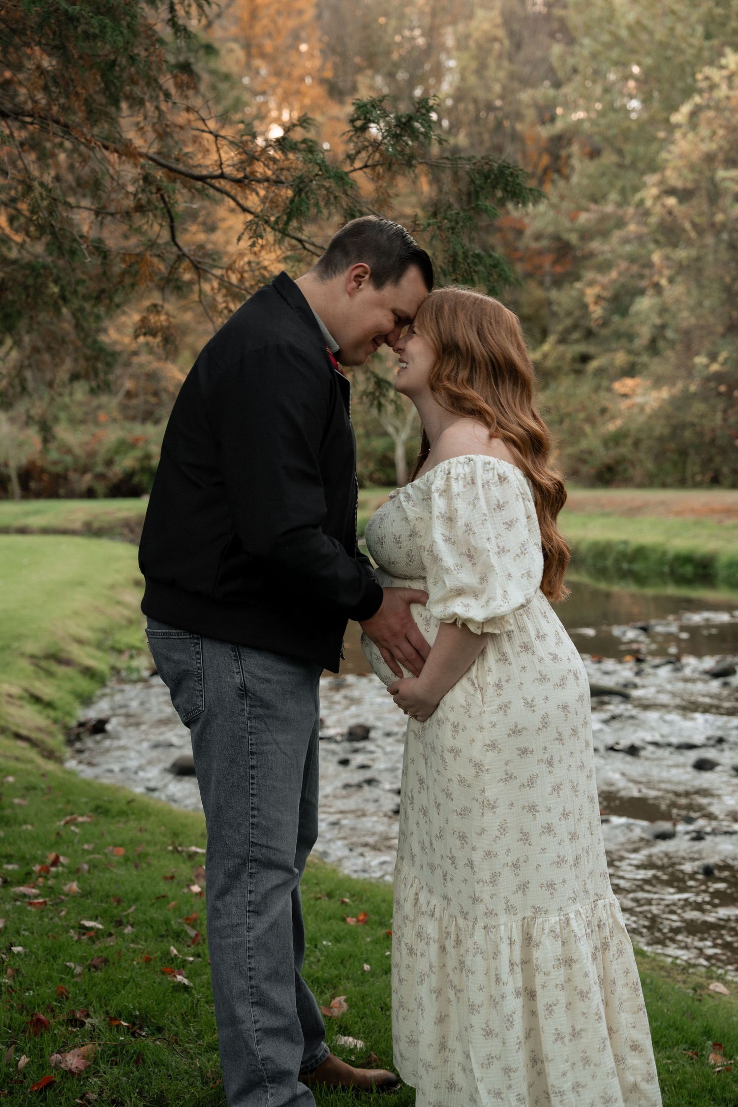 Pregnant woman in white floral dress standing in a serene forest pond at sunset during autumn.