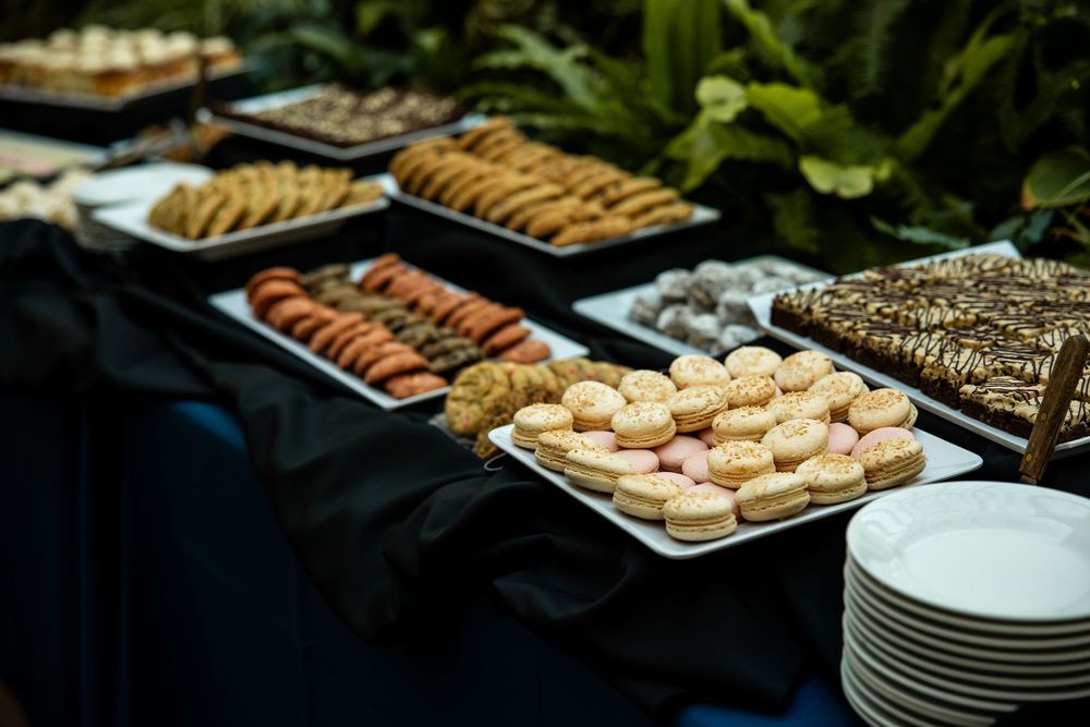 Elegant dessert buffet table with macarons, cookies, and pastries displayed on trays with stacked white plates nearby.