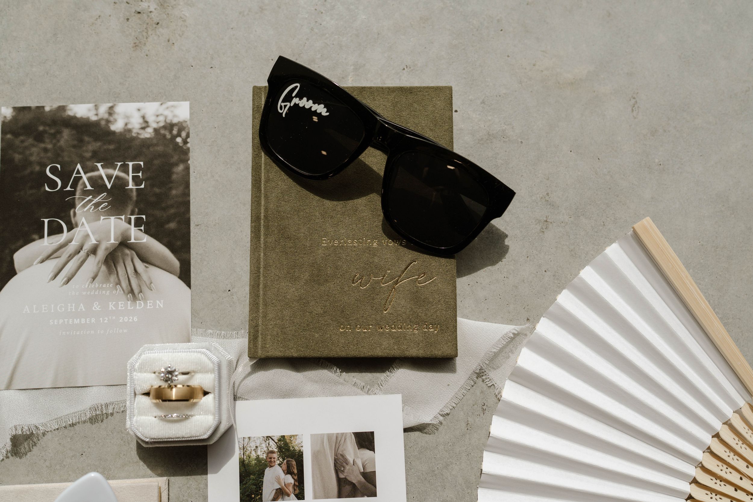 Wedding flat lay with save the date card, sunglasses, ring box, fan, and photos on gray surface.