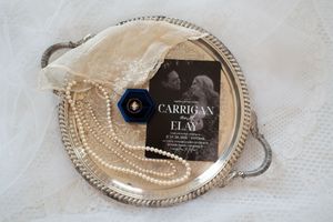 Silver tray with pearl necklace, blue ring, lace veil, and Carrigan & Elay wedding invitation flat lay.