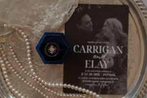 Wedding flat lay featuring a Carrigan and Elay invitation, pearl necklace, and a navy hexagonal ring box on a silver tray.