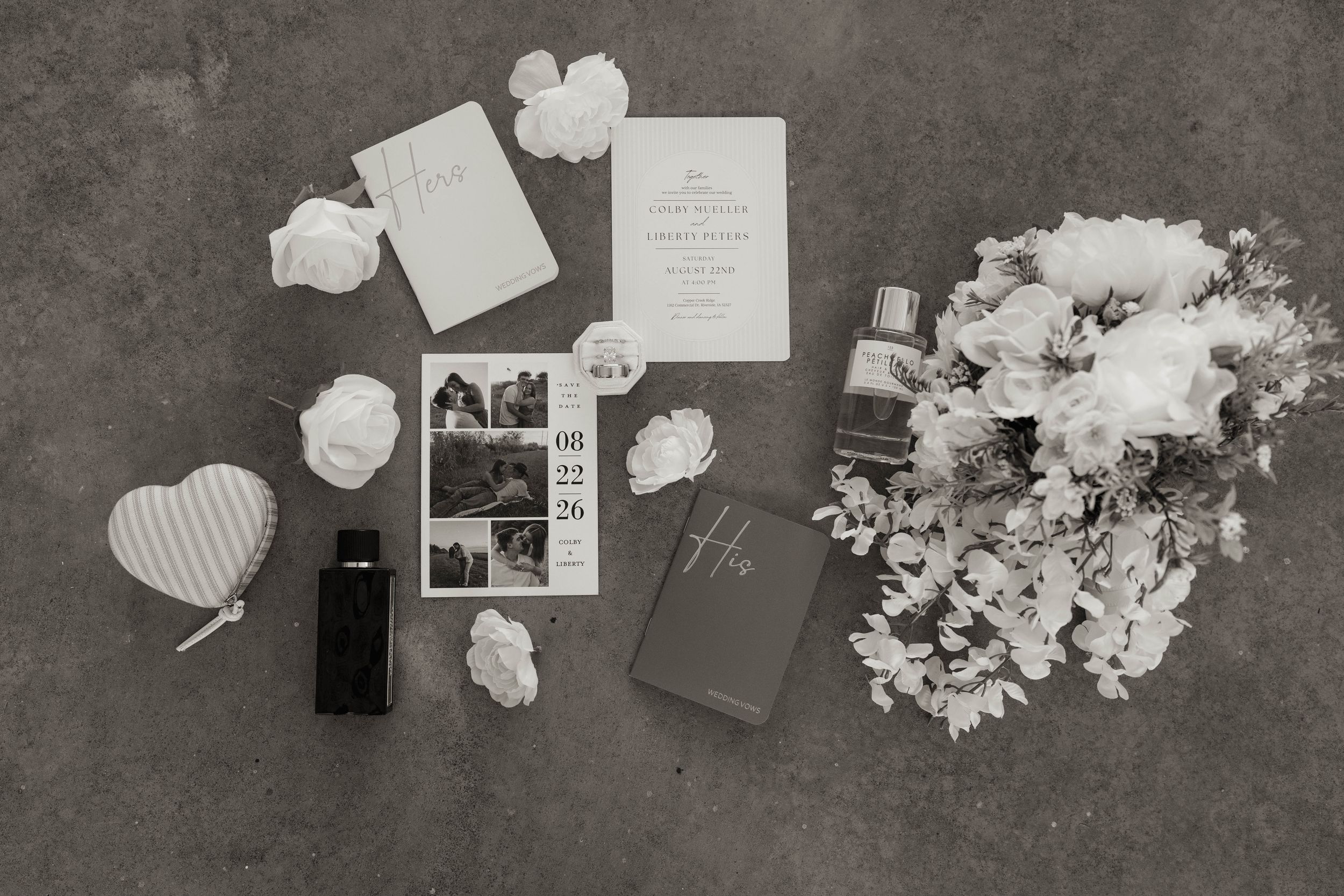 Black and white flat lay of wedding stationery, bouquet, perfume, and polaroid photos on a gray surface.