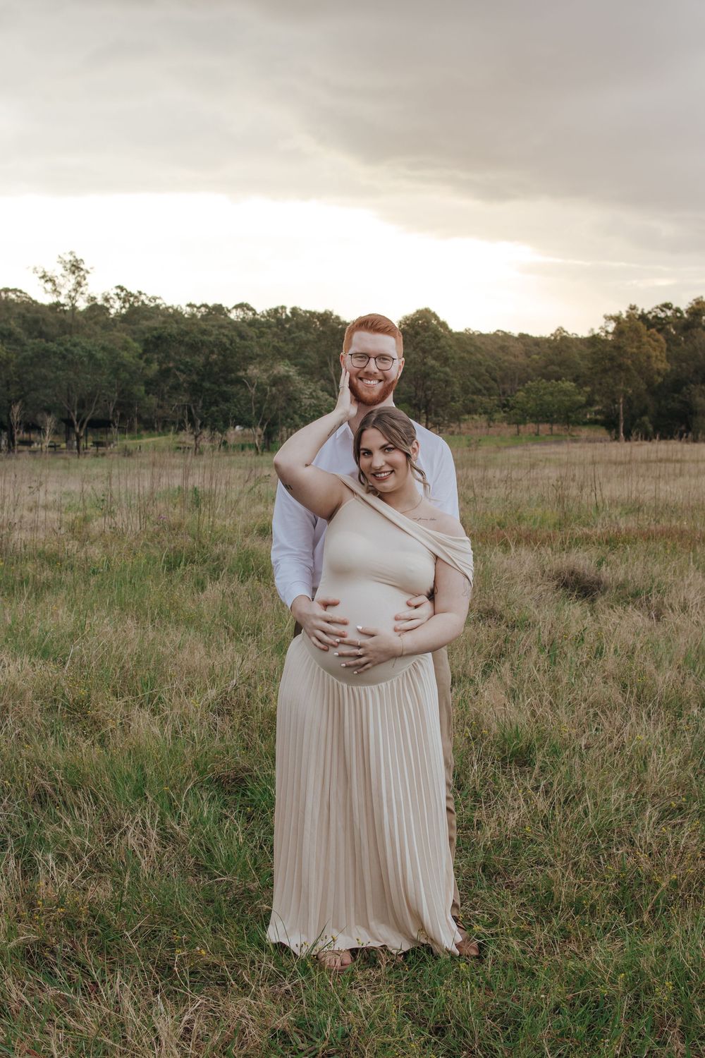 Couple posing in a field at sunset in Rouse Hill, woman in beige pleated maternity dress cradling baby bump, man embracing her from behind.