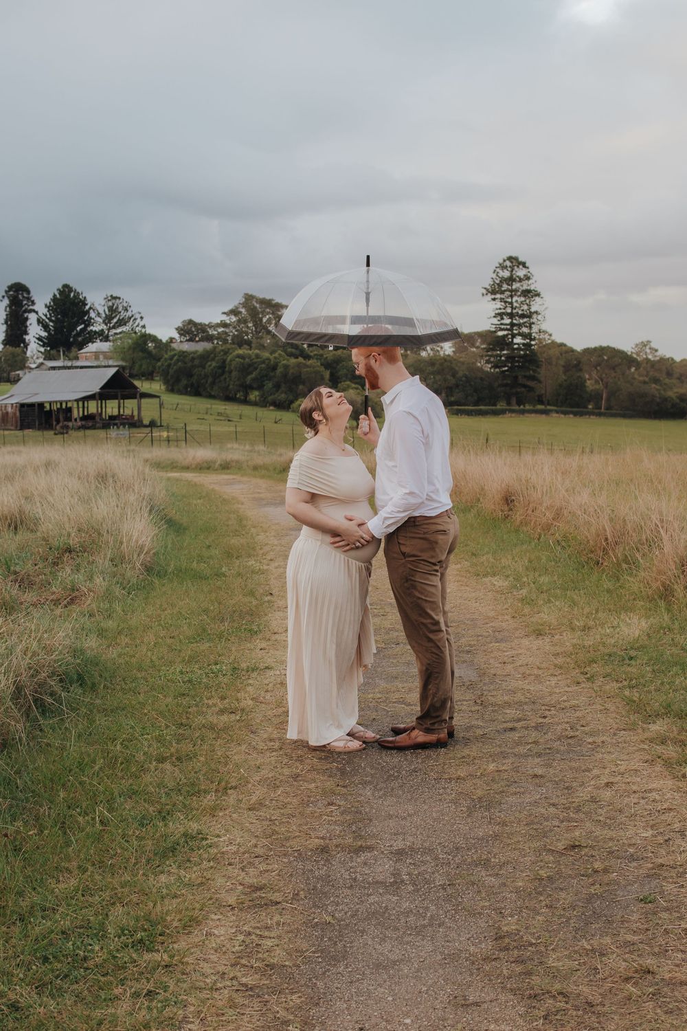 Pregnant couple sharing a tender moment under a clear umbrella on a rural path surrounded by golden grasslands.