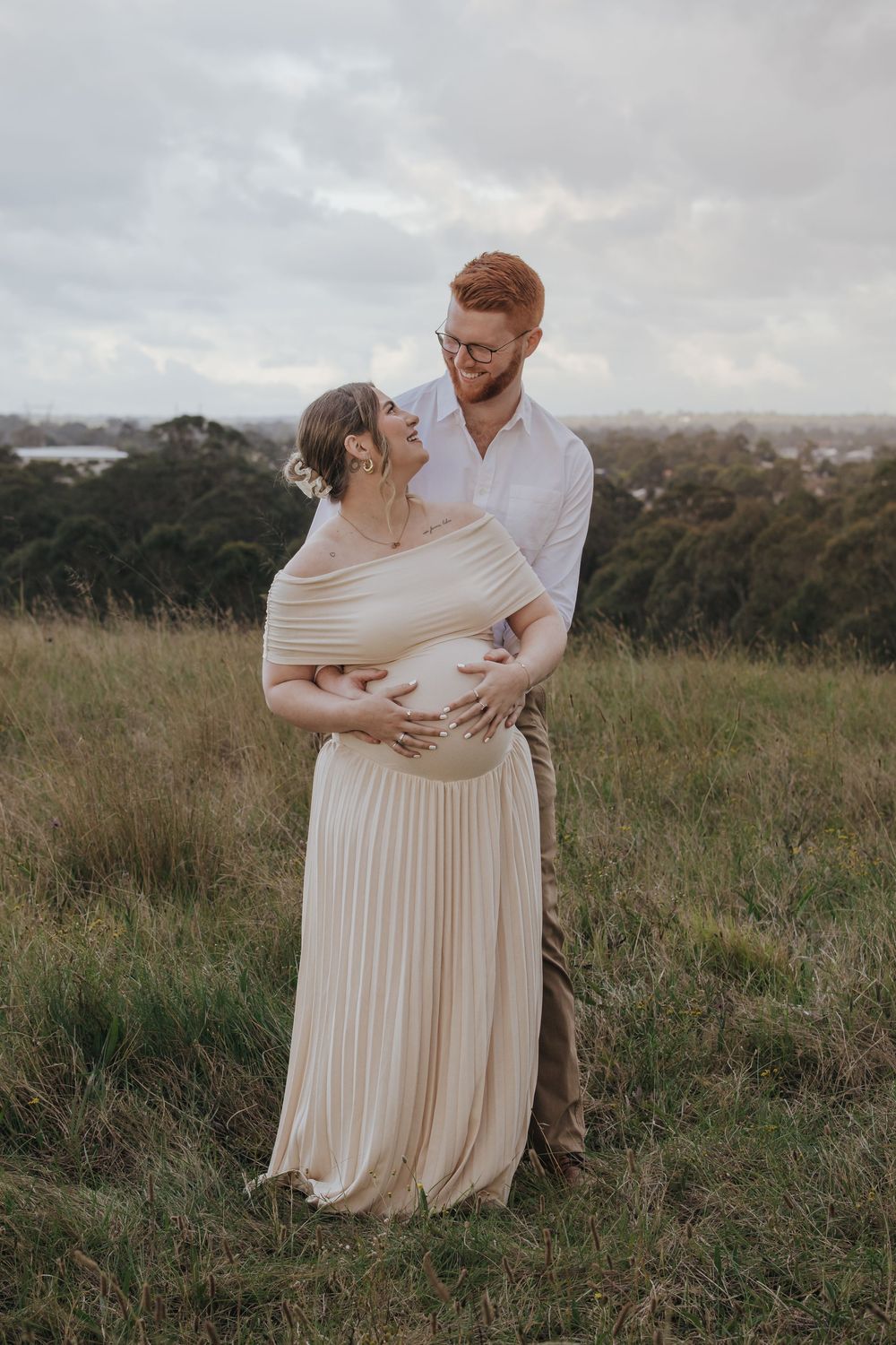 Couple sharing a tender moment outdoors, pregnant woman in cream pleated dress, man in white shirt, lush green hillside backdrop.