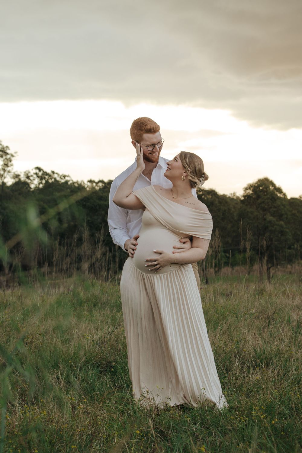 Couple sharing a tender moment in a field at sunset, pregnant woman in cream pleated gown cradling her baby bump.