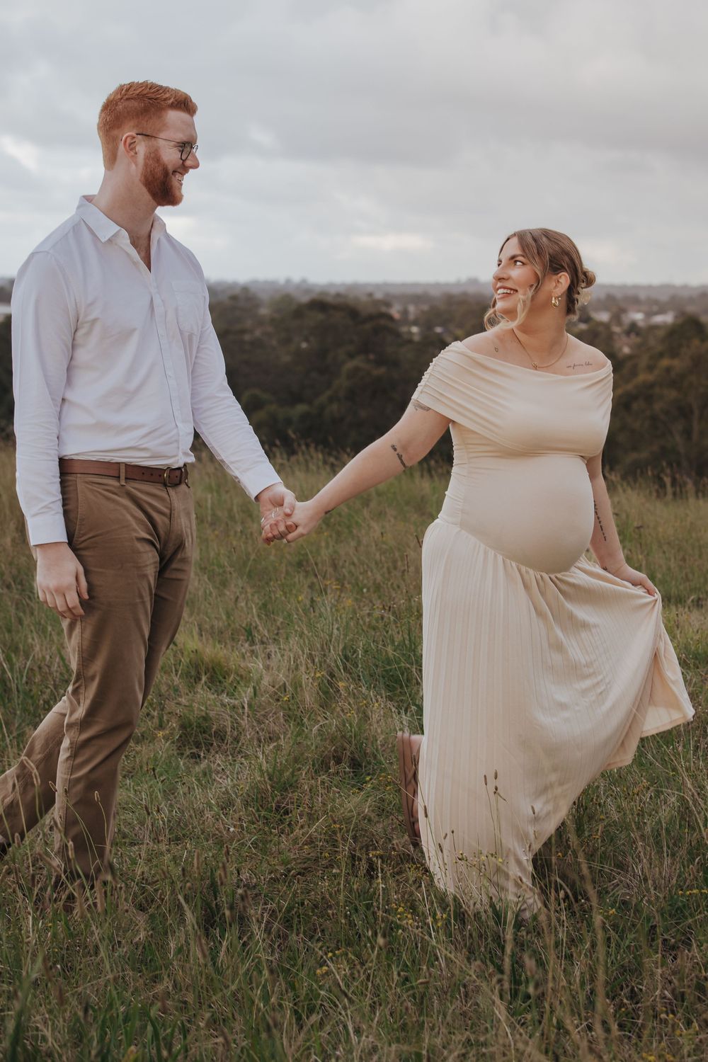 Pregnant couple holding hands and smiling while walking through a green field, woman in cream dress, man in white shirt.