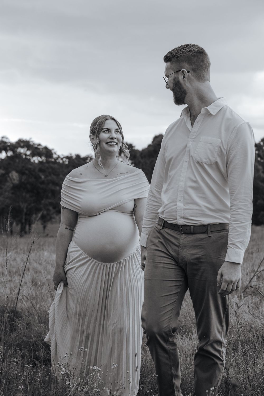 Black and white photo of a smiling pregnant woman in a flowing dress with her partner in a field.