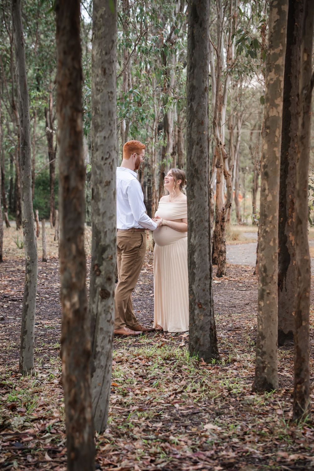 Pregnant couple holding hands in a sunlit eucalyptus forest, woman in cream dress, man in white shirt at Rouse Hill Regional Park.