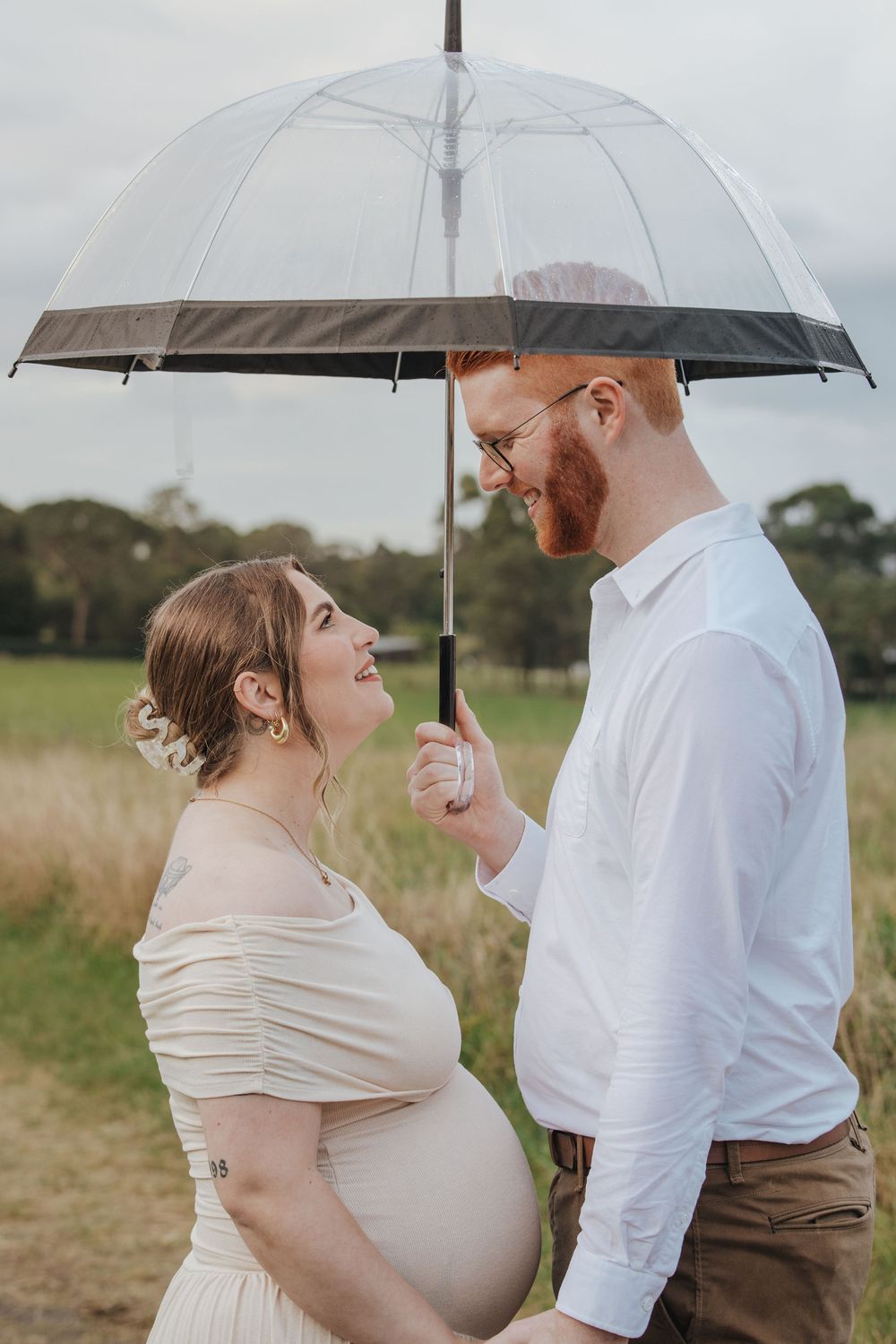 Pregnant couple smiling at each other under a clear umbrella outdoors in a green field, maternity photo session.