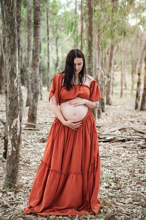 Pregnant woman in rust red boho dress cradling baby bump in a serene forest setting at Rouse Hill Regional Park.