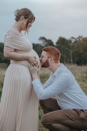 A man kneels kissing a pregnant woman's belly in a field in Rouse Hill, capturing a tender maternity moment outdoors.