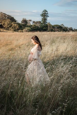 Pregnant woman in white lace dress standing in golden field at sunset, cradling baby bump.