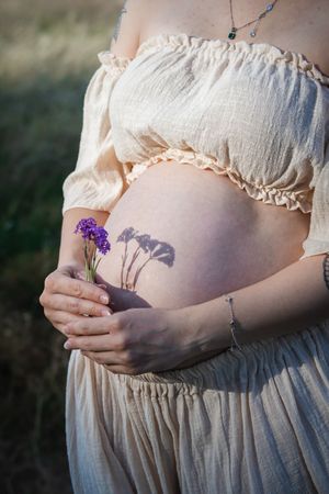 Pregnant woman in cream dress holding purple wildflowers against her bare belly outdoors in soft golden light.