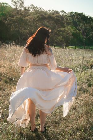 Woman in flowing peach off-shoulder dress walking through sunlit meadow with trees in background in Rouse Hill.