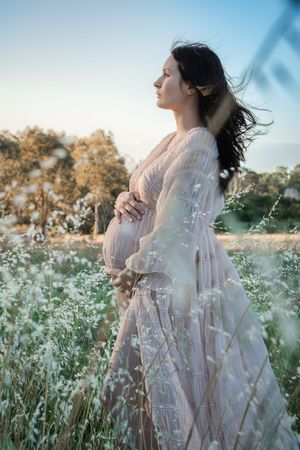 Pregnant woman in flowy dress standing in wildflower field, cradling belly at golden hour.