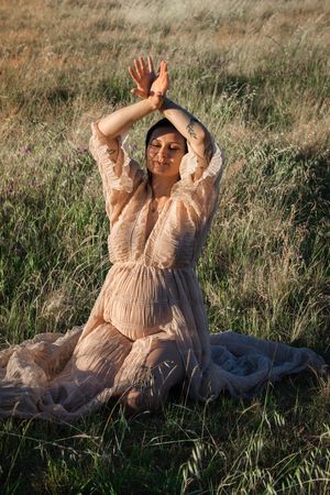 A woman in a flowing beige dress sits in a grassy field with arms raised gracefully overhead in golden sunlight.
