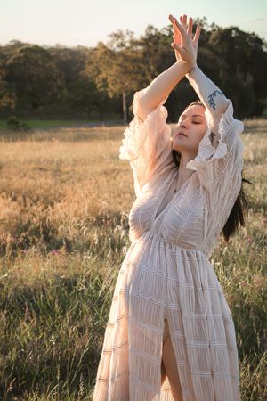 Woman in white lace dress standing in golden field with arms raised, bathed in warm sunset light.
