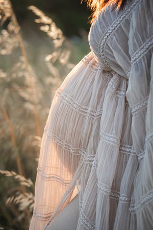 Pregnant woman in white lace boho dress standing in golden field at Rouse Hill Regional Park at sunset, showcasing baby bump.