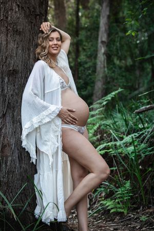 Smiling pregnant woman in white kimono and lingerie posing against a tree in a lush green forest.
