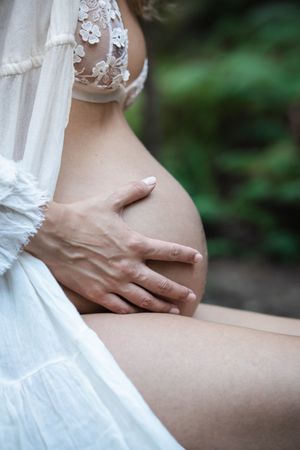 Pregnant woman in white lace dress gently cradling her baby bump outdoors with green foliage background.