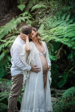 Couple sharing a tender kiss in a fern forest, man embracing pregnant woman in white lace maternity robe.