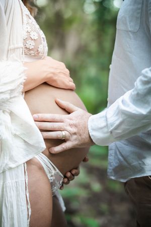 Couple tenderly cradling pregnant belly outdoors, woman in white lace dress, man in light blue shirt, nature background.