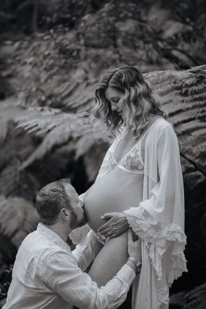 Black and white photo of man kneeling to kiss pregnant woman's belly in a lush fern forest.