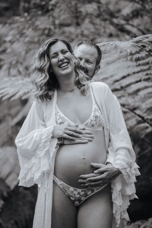 Black and white photo of a laughing pregnant woman in white robe and lingerie embraced by partner outdoors.