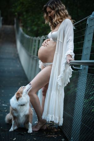 Pregnant woman in white robe and lingerie on the Swing Bridge in Cherrybrook with a small white dog looking up at her belly.