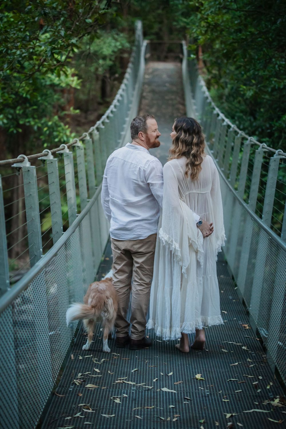 Couple in white attire standing on Swing Bridge in Cherrybrook with their golden dog, sharing a romantic moment.