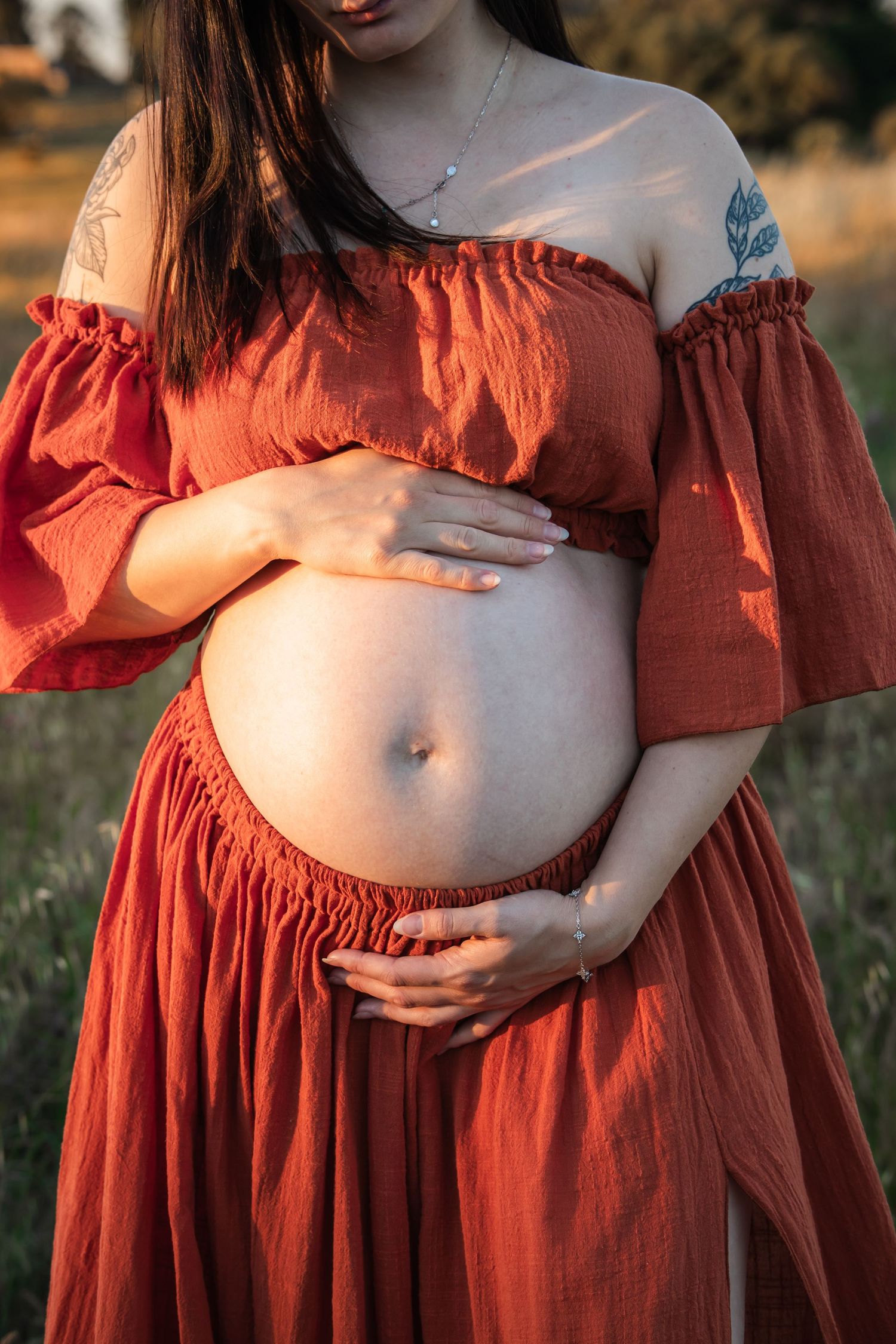 Pregnant woman in red off-shoulder dress cradling her bare baby bump outdoors in golden sunlight in Maternity photoshoot in Rouse Hill.