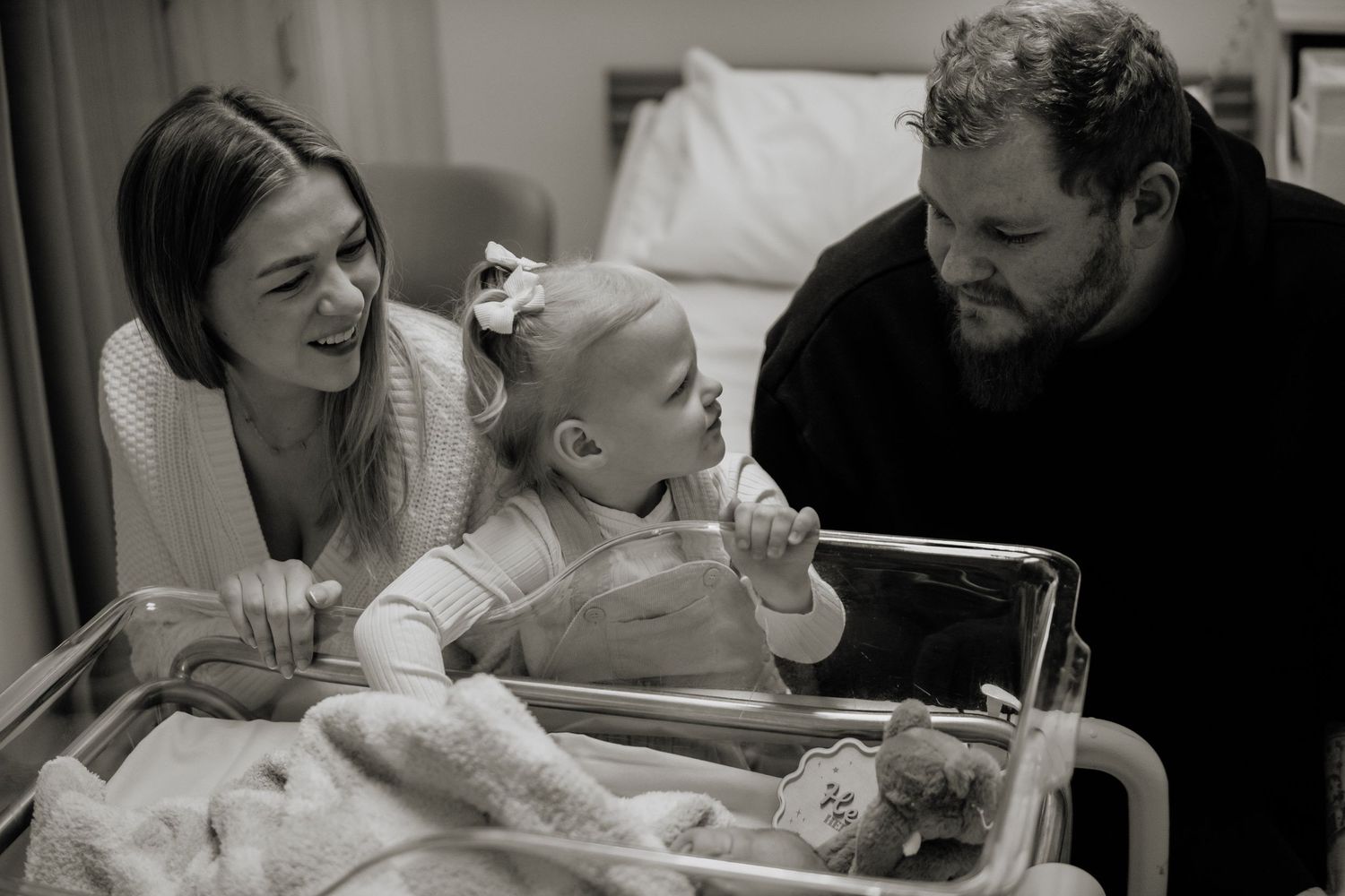 Black and white photo of parents smiling at toddler beside hospital newborn bassinet during fresh 48 photoshoot in the Hawkesbury.